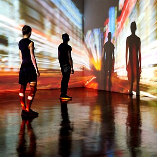 In einem abgedunkelten Raum stehen zwei Personen auf einem glänzenden Boden und betrachten eine großflächige Projektion an den Wänden. Die Projektion zeigt dynamische, stark verwischte Lichtspuren und Farbstreifen, die an schnelle Bewegung in einer Großstadt bei Nacht erinnern. Die Personen sind als dunkle Silhouetten vor den leuchtenden Hintergründen sichtbar, und ihre Schatten werden groß und deutlich auf die Projektion geworfen. Die Szene wirkt immersiv, mit Lichtreflexen auf dem Boden und intensiven Farbverläufen an den Wänden.