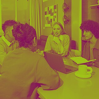 Varias personas están sentadas alrededor de una mesa en un espacio interior, conversan entre sí y tienen una computadora portátil, una taza y un cuaderno abierto sobre la mesa.