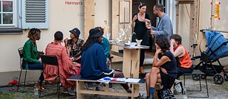 Eine Gruppe von Personen sitzt im Freien in einem halbkreisförmigen Arrangement vor einem Gebäude mit hellen Wänden und geöffneten Türen. Die Personen sitzen auf verschiedenen Stühlen und Hockern um einen niedrigen Tisch, auf dem Papier, Stifte und weitere Materialien liegen. Rechts daneben steht ein höherer Stehtisch mit Getränke‑ und Snackbehältern sowie einem dekorativen Objekt aus dünnen Stäben. Im Hintergrund befindet sich ein Kinderwagen. Einige Personen stehen, andere sitzen, während sie sich unterhalten oder Materialien in den Händen halten. An der Wand des Gebäudes ist der Schriftzug „Herman’s“ zu sehen.