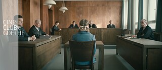 Ein Mädchen mit geflochtenen Zöpfen sitzt in einem Gerichtssaal im Zeugenstand. Sie ist nur von hinten zu sehen. Sie wird von älteren Richtern kritisch angeschaut.