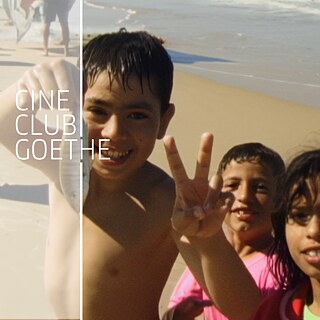 Drei Kinder spielen am Strand. Sie tragen Badekleidung und lachen in die Kamera. Eines der Kinder hält einen kleinen Fisch hoch und zeigt mit seiner Hand das "Peace"-Zeichen.