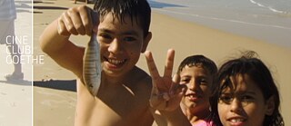 Drei Kinder spielen am Strand. Sie tragen Badekleidung und lachen in die Kamera. Eines der Kinder hält einen kleinen Fisch hoch und zeigt mit seiner Hand das "Peace"-Zeichen.