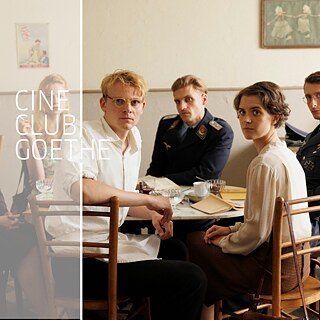 Eine Gruppe von sechs jungen Erwachsenen sitzt an einem kleinen Cafétisch und blickt direkt in die Kamera, mit ernsten und aufmerksamen Gesichtsausdrücken. Die Umgebung wirkt schlicht und sanft beleuchtet, mit Holzstühlen sowie Tassen, Gläsern und Papieren auf dem Tisch. Zwei Männer auf der rechten Seite tragen militärisch wirkende Uniformen, während die anderen in legerer Kleidung aus der Mitte des 20. Jahrhunderts gekleidet sind. Eine Frau hält eine Zigarette. Die Stimmung erscheint insgesamt angespannt und nachdenklich. Auf der linken Seite des Bildes steht der Text „CINE CLUB GOETHE“.