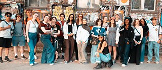  Eine Gruppe junger Teilnehmender steht gemeinsam vor einer urbanen Backsteinwand mit Plakaten und Graffiti und posiert im Rahmen eines internationalen Austauschprojekts.