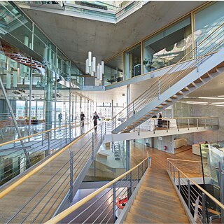 Ein großer, moderner Innenraum mit mehreren Ebenen, Glaswänden und offenen Treppenanlagen. Ein weitläufiges Netz aus Metallgeländern, Holzstegen und schwebenden Treppen verbindet verschiedene Stockwerke. Im Zentrum verlaufen breite Treppen und Rampen mit Holzbelag. Auf der linken Seite des Bildes spiegeln große Glasflächen den Innenraum und zeigen dahinterliegende Arbeits- oder Aufenthaltsbereiche mit Tischen und Stühlen. An der Decke hängen markante, vertikal angeordnete Leuchten. Im oberen Bereich fallen Tageslichtöffnungen und große Fenster ins Gebäude. Einige Personen bewegen sich auf den Wegen und Treppen durch den Raum.