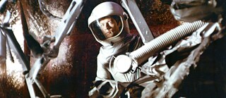 Persona con traje espacial y casco transparente, rodeada de tubos técnicos y estructuras metálicas. La escena está iluminada de forma tenue y recuerda a un entorno de ciencia ficción.