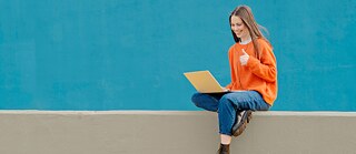 Persona sentada sobre un muro bajo frente a una pared de color turquesa, utilizando una computadora portátil.