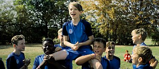 Un grupo de jóvenes futbolistas con camisetas azules se encuentra en un campo de césped. Varias personas del equipo levantan a una sobre sus hombros; al fondo se observan árboles y otra cancha.
