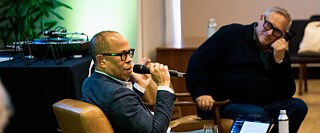 Michael Bowie speaks into a microphone while seated in the foreground during the Disko Diskurs event. In the background, Thomas Meinecke listens while holding a microphone. A turntable setup and event equipment are visible beside them.