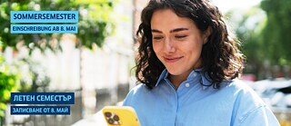 Млада жена на улицата се усмихва, гледайки телефона си.