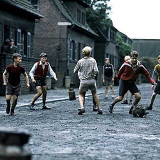 Escena histórica al aire libre: Un grupo de niños juega con una pelota en una calle sin pavimentar entre edificios de ladrillo. Los niños llevan ropa sencilla, están en movimiento y distribuidos por la calle, con más construcciones visibles al fondo.