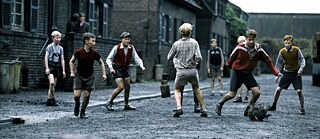 Escena histórica al aire libre: Un grupo de niños juega con una pelota en una calle sin pavimentar entre edificios de ladrillo. Los niños llevan ropa sencilla, están en movimiento y distribuidos por la calle, con más construcciones visibles al fondo.