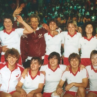 Fotografía histórica de un equipo deportivo femenino: Varias personas con camisetas blancas y pantalones rojos posan juntas en varias filas, de pie y agachadas. La imagen fue tomada en un estadio, con público visible al fondo.