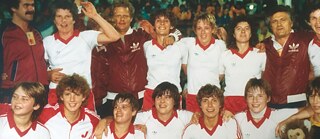 otografía histórica de un equipo femenino deportivo: Varias personas con camisetas blancas y pantalones rojos posan juntas en varias filas, de pie y agachadas. La imagen fue tomada en un estadio, con público visible al fondo.