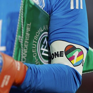 Primer plano de un futbolista con camiseta azul y guantes de arquero. En el brazo lleva un brazalete multicolor “One Love”, junto a un banderín verde relacionado con el fútbol alemán.