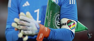 Primer plano de un futbolista con camiseta azul y guantes de arquero. En el brazo lleva un brazalete multicolor “One Love”, junto a un banderín verde relacionado con el fútbol alemán.
