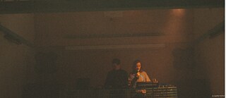 In einem großen, abgedunkelten Raum stehen zwei Menschen nebeneinander hinter einer erhöhten, gitterartigen Installation, die an ein technisches oder klangbasiertes Gerät erinnert. Warmes, gedämpftes Licht und leichter Nebel umgeben die beiden Personen und verleihen der Szene eine ruhige, atmosphärische Wirkung. 