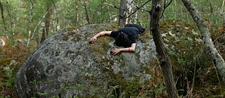 Eine Person liegt bäuchlings über einem großen, moosbedeckten Felsblock in einem dichten Wald. Die Arme hängen seitlich über den Stein herab, umgeben von Bäumen, Farnen und Waldboden. Das Foto zeigt eine ruhige Waldszene mit natürlichen Farben und weichem Licht. 
