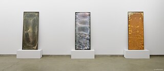 In einem hellen Ausstellungsraum stehen drei schmale, hochformatige Tafeln auf weißen Sockeln nebeneinander an einer weißen Wand. Jedes Objekt zeigt eine unterschiedliche, reliefartige Oberfläche mit variierenden Farben und Texturen. Die klare Präsentation und das gleichmäßige Licht lenken die Aufmerksamkeit auf Materialität und Struktur der Werke. 