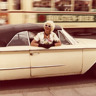 Eine Frau mit blonden Locken und Sonnenbrille lehnt lächelnd aus dem Fenster eines hellen Cabriolets, während die Stadt im Hintergrund verschwimmt. Die Szene wirkt dynamisch und nostalgisch, als wäre sie in einer früheren Zeit aufgenommen worden. 