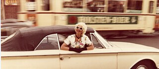 Eine Frau mit blonden Locken und Sonnenbrille lehnt lächelnd aus dem Fenster eines hellen Cabriolets, während die Stadt im Hintergrund verschwimmt. Die Szene wirkt dynamisch und nostalgisch, als wäre sie in einer früheren Zeit aufgenommen worden. 