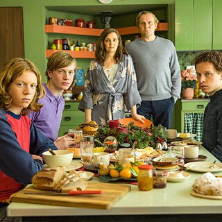 Una familia de cinco personas están sentadas y de pie alrededor de una mesa de comedor en una cocina de color verde. Sobre la mesa hay pan, fiambres, queso, vajilla y alimentos; al fondo se ven armarios y estanterías de cocina.