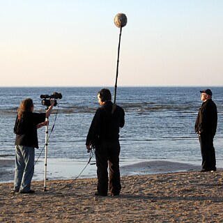 Drei Personen am Strand beim Filmdreh. Links mit Kamera, in der Mitte mit Tonangel und rechts der Regisseur. 