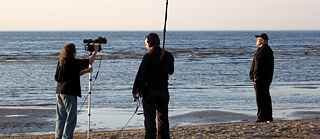 Drei Personen am Strand beim Filmdreh. Links mit Kamera, in der Mitte mit Tonangel und rechts der Regisseur. 