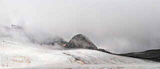 Ein Gletscher mit rötlich-braunem Staub im Vordergrund und nebelverhangenen Berggipfeln im Hintergrund.