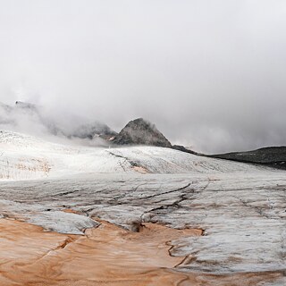 Ein Gletscher mit rötlich-braunem Staub im Vordergrund und nebelverhangenen Berggipfeln im Hintergrund.