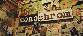 Ein dicht beklebter Innenraum zeigt eine grüne Wand voller Collagen, Plakate, Zeichnungen und humorvoller Schilder, darunter ein großes Schild mit der Aufschrift „monochrom“. 