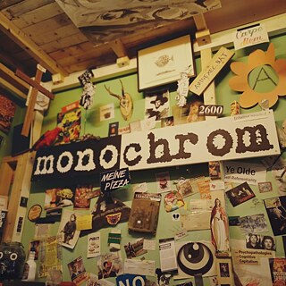 Ein dicht beklebter Innenraum zeigt eine grüne Wand voller Collagen, Plakate, Zeichnungen und humorvoller Schilder, darunter ein großes Schild mit der Aufschrift „monochrom“. 