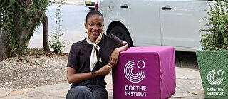 Ndayola Ulenga sitzt auf einer Treppenstufe, neben ihr ein lilafarbener Stoffwürfel mit dem Logo des Goethe-Instituts