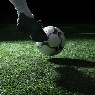Un balón de fútbol sobre césped artificial verde, presionado suavemente por un botín deportivo. La escena está iluminada de forma tenue, evocando un estadio de noche.