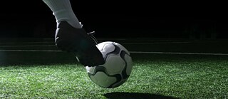 Un balón de fútbol sobre césped artificial verde, presionado suavemente por un botín deportivo. La escena está iluminada de forma tenue, evocando un estadio de noche.