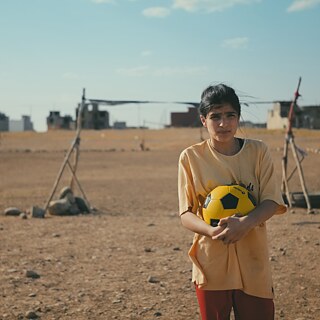 Una joven sostiene un balón de fútbol amarillo y negro y está de pie en un campo abierto y polvoriento. Al fondo se observan porterías improvisadas, piedras esparcidas y edificios a lo lejos.
