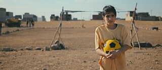 Una joven sostiene un balón de fútbol amarillo y negro y está de pie en un campo abierto y polvoriento. Al fondo se observan porterías improvisadas, piedras esparcidas y edificios a lo lejos.