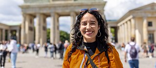 Eine Deutschschülerin lächelt strahlend in die Kamera. In ihrem orangefarbenen Outfit strahlt sie Stolz und Freude aus. Sie steht am Pariser Platz in Berlin, nahe dem Brandenburger Tor. Die beeindruckende Kulisse unterstreicht die besondere Stimmung des Moments.