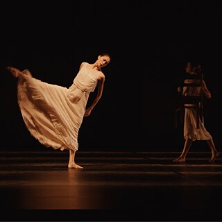 Persona baila con un vestido blanco fluido en un escenario oscuro, mientras otra persona sostiene una silla al fondo.