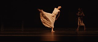 Persona baila con un vestido blanco fluido en un escenario oscuro, mientras otra persona sostiene una silla al fondo.