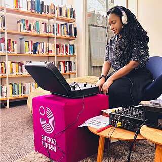  Person wearing headphones operates a record player at a listening station in a library, surrounded by shelves of books and audio equipment.