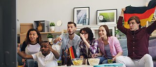 A group of people sitting on a couch watching TV with a German flag. Popcorn and chips are on the TV table.