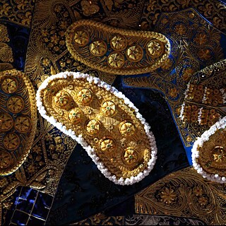 Detailaufnahme mehrerer aufwendig verzierter Objekte aus goldenem Material auf dunkelblauem Stoff. Die länglichen Formen sind mit runden, erhabenen Mustern, feinen Stickereien und kleinen weißen Perlen entlang der Ränder besetzt. Im Hintergrund sind weitere textile Elemente mit dichten, ornamentalen Verzierungen sichtbar. Die Oberfläche zeigt eine Mischung aus glänzenden und matten Strukturen.