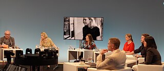 Members of the jury sit in a semi-circle on white armchairs, all looking uniformly to the left. In the background, Ingeborg Bachmann is shown in black and white on a screen.