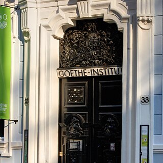 Vchod do historickej budovy s ozdobnými dverami, nadpis „GOETHE-INSTITUT“. Svetlá fasáda, číslo 33 viditeľné, zelený banner vľavo.
