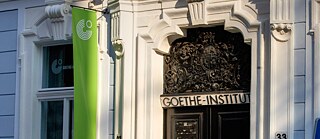 Eingang eines historischen Gebäudes mit verzierter Tür, über dem „GOETHE-INSTITUT“ steht. Helle Fassade, Nummer 33 sichtbar, grünes Banner links.