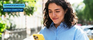 Eine junge Frau steht auf dem Gehweg, lächelt und schaut auf ihr Handy.