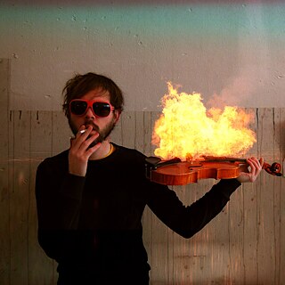 Un hombre con gafas de sol rojas sostiene un violín en llamas mientras fuma un cigarrillo; al fondo se ve una pared y una imagen de una explosión.