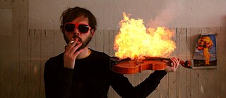 Un hombre con gafas de sol rojas sostiene un violín en llamas mientras fuma un cigarrillo; al fondo se ve una pared y una imagen de una explosión.