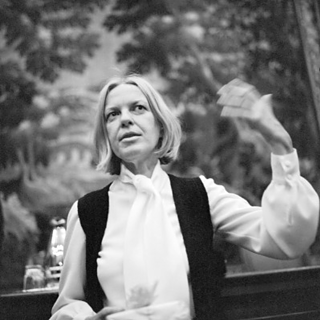 Auf einem Schwarz-Weiß-Foto ist die österreichische Schriftstellerin Ingeborg Bachmann zu sein, einen Mittelscheitel tragend, ihre linke Hand ist durch eine gestikulierende Bewegung nur verschwommen zu sehen.